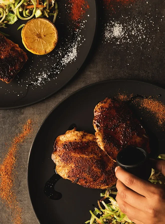 Hand pours sauce onto grilled chicken and salad on dark plates.