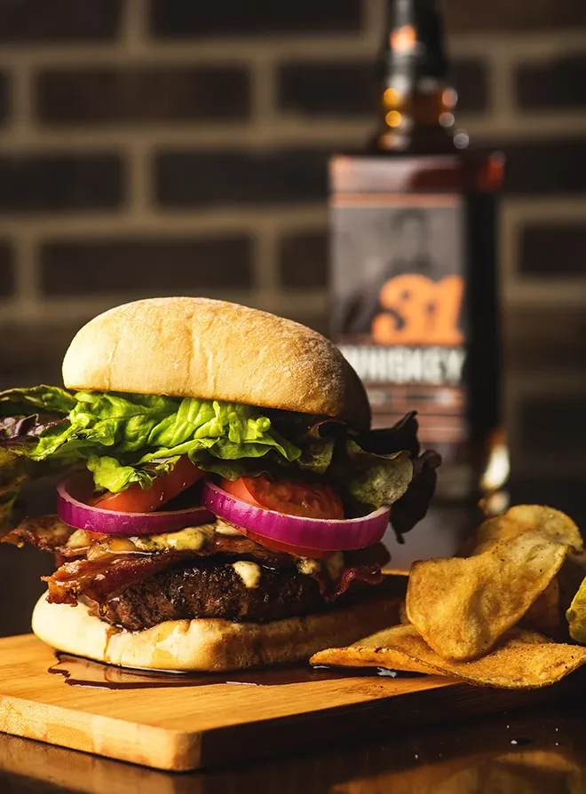 Large burger with bacon, lettuce, tomato, chips, and whiskey bottle.