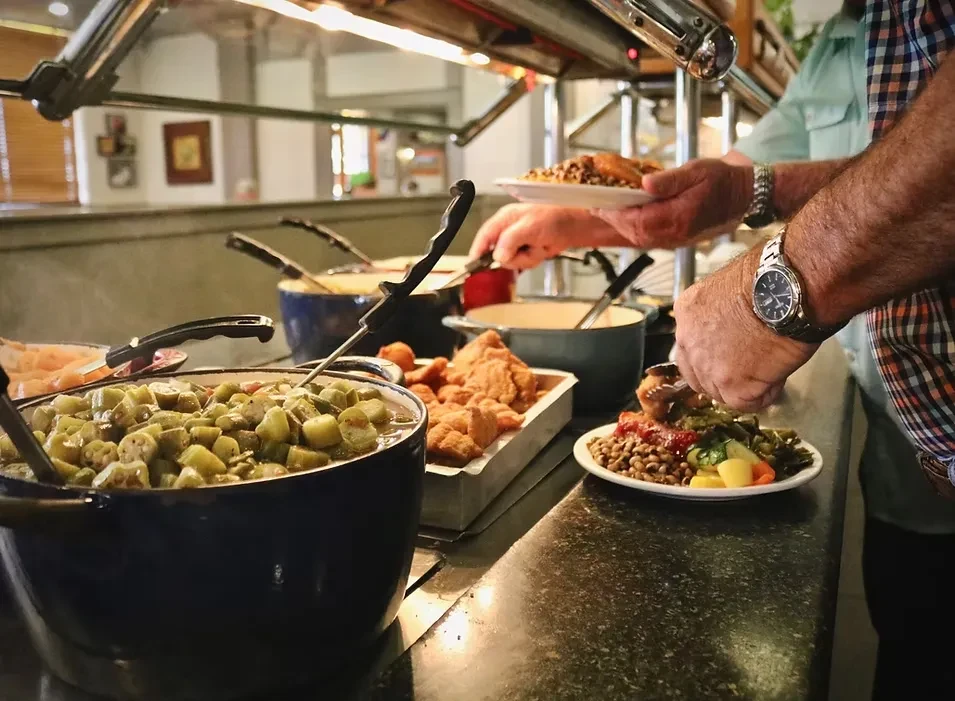 Hands serving okra stew and fried chicken at a buffet line.