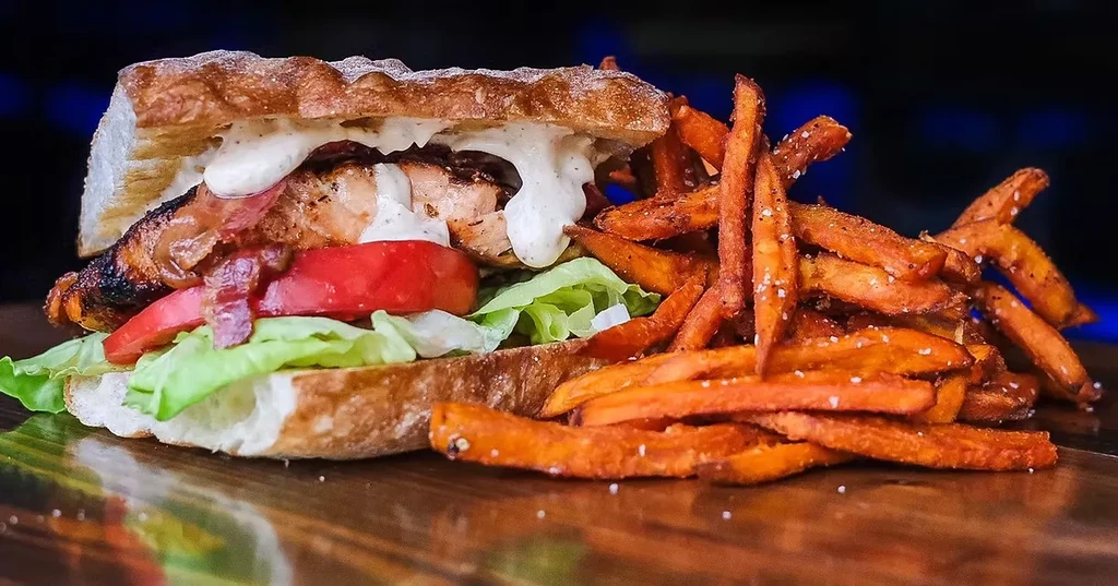Grilled sandwich with meat, bacon, vegetables, and seasoned sweet potato fries.