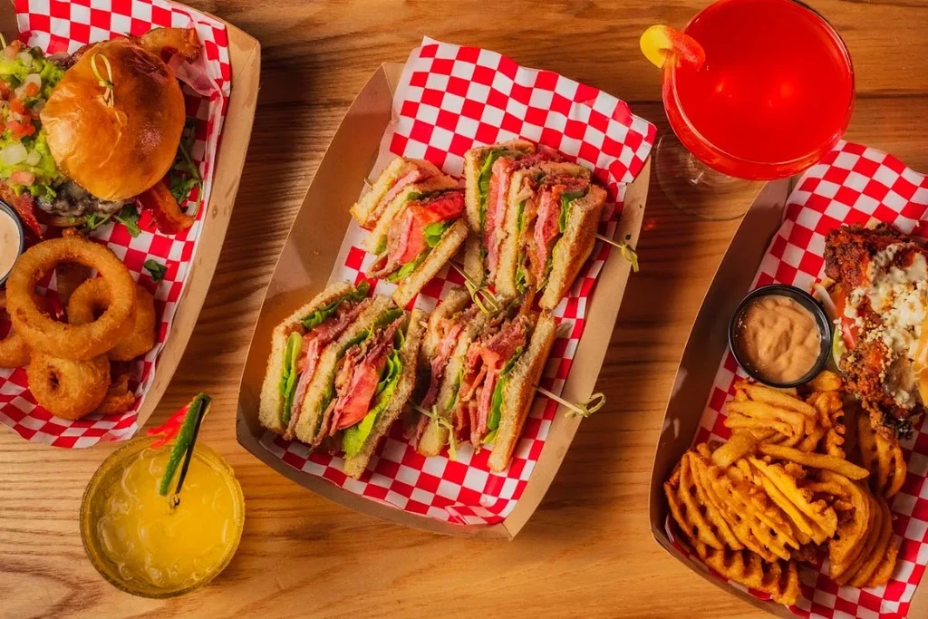 Three trays with a burger, sandwiches, fries, and two cocktails.