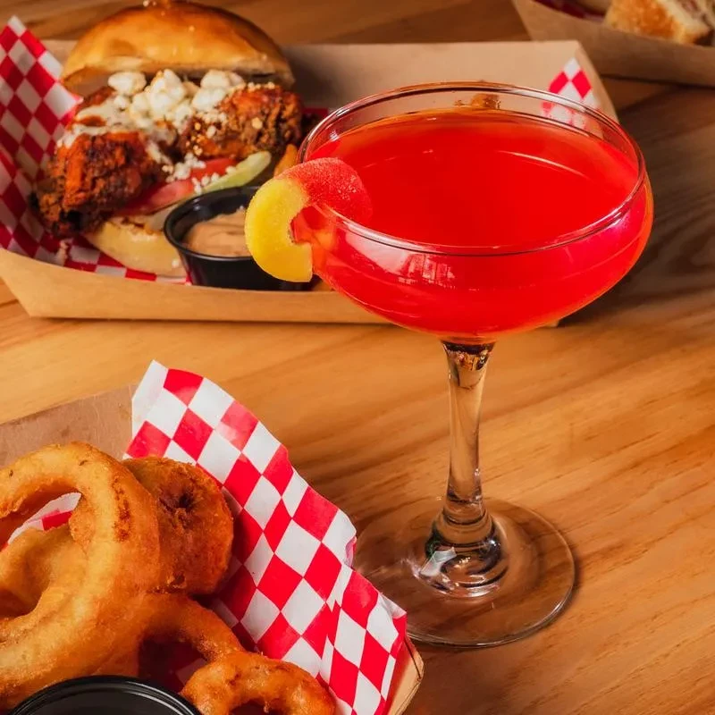Red cocktail, fried chicken sandwich, and onion rings on a wooden table.