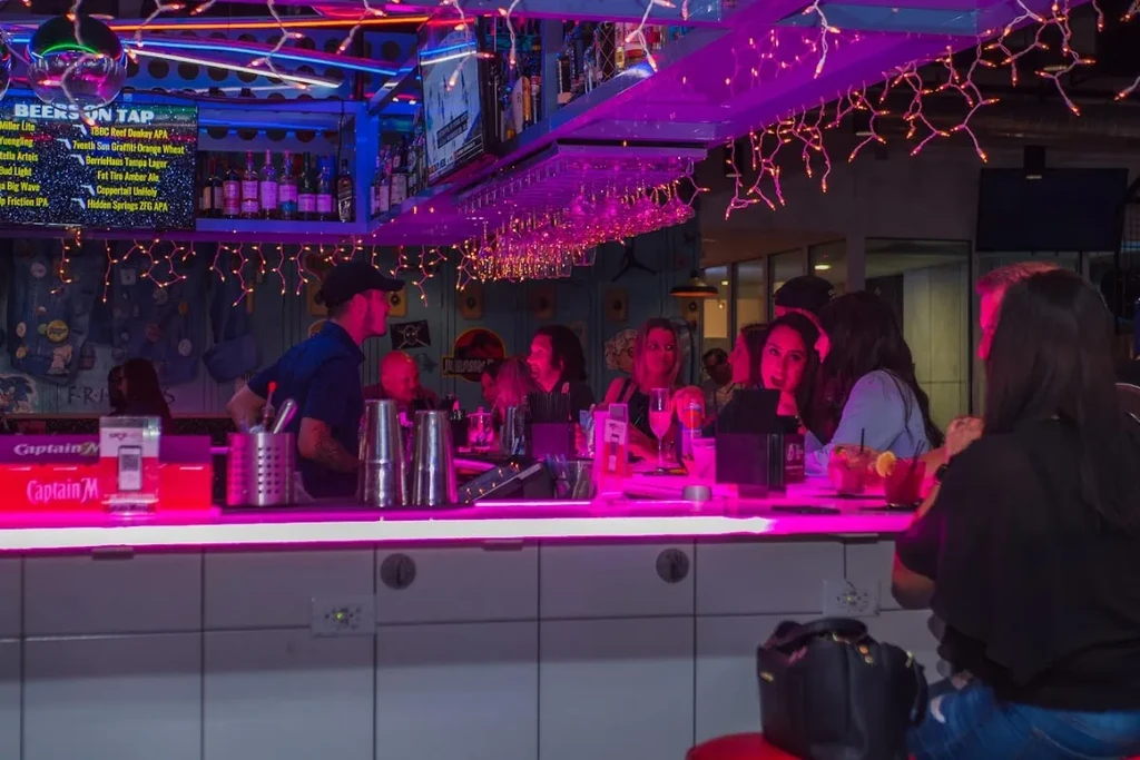 Lively, neon-lit bar scene with patrons and a bartender.