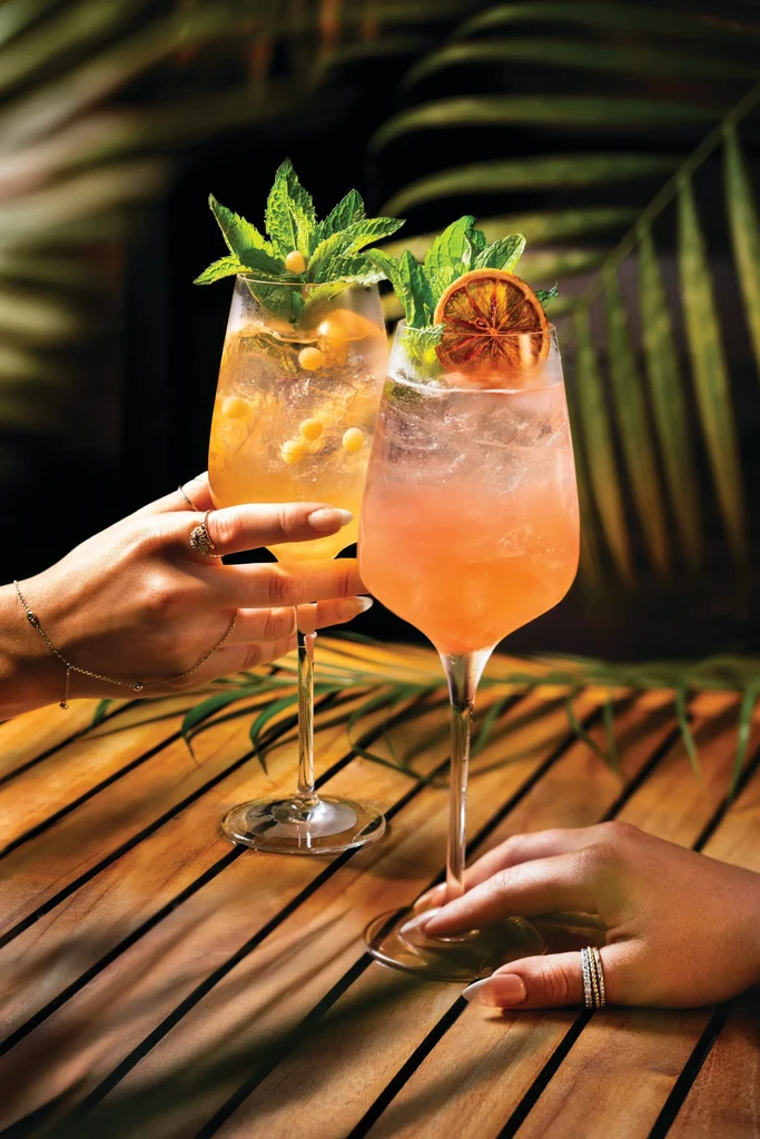 Hands clinking two colorful, garnished cocktails in a tropical setting.