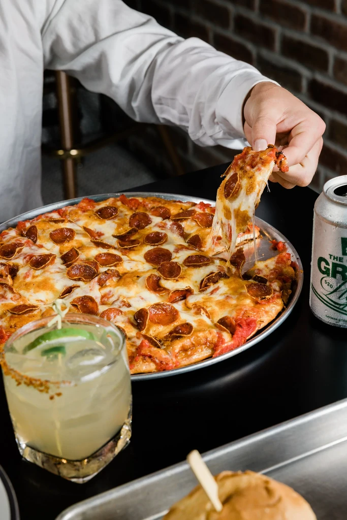 Hand pulls cheesy pepperoni pizza slice; cocktail, beer, burger nearby.