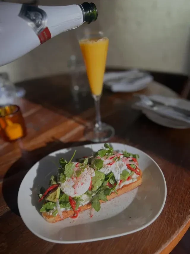 Avocado toast with poached eggs, greens, and a mimosa pouring.