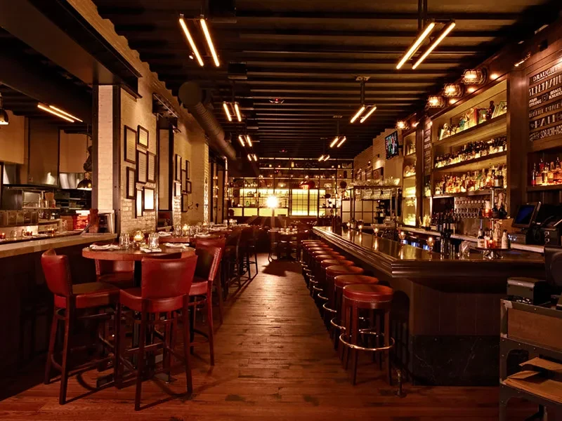 Dimly lit, warm restaurant and bar interior with wooden bar and red stools.