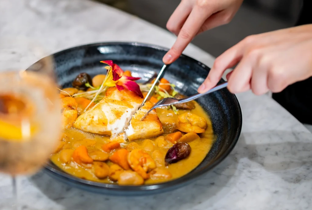 Hands cutting pan-seared white fish with shrimp, vegetables, and orchid.