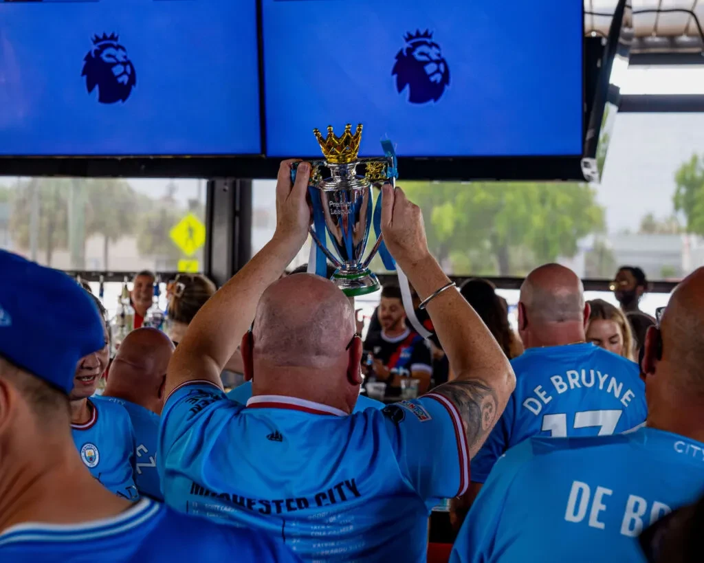 Manchester City fan raises replica Premier League trophy amidst celebrating fans.