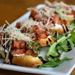 Bruschetta topped with tomatoes, cheese, basil, balsamic, and arugula garnish.