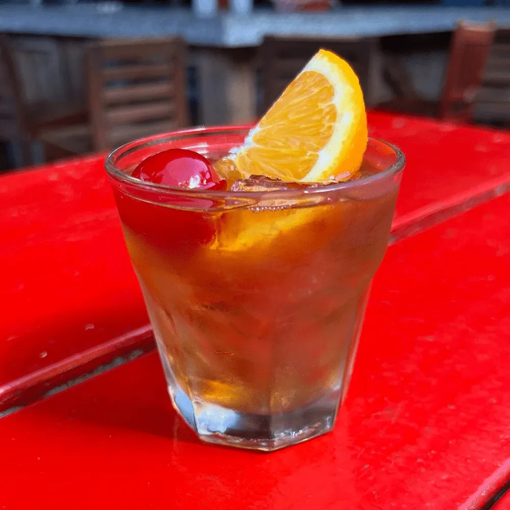 Amber cocktail with cherry and orange in faceted glass on red table.