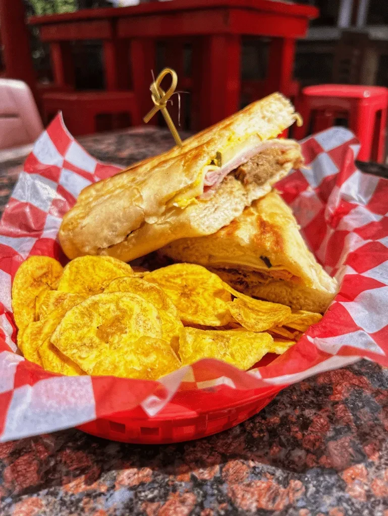Halved Cuban-style pressed sandwich with plantain chips in a red basket.