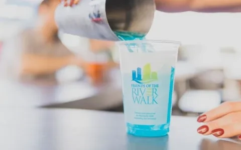 A hand pours blue liquid into a "FRIENDS OF THE RIVER WALK" cup.