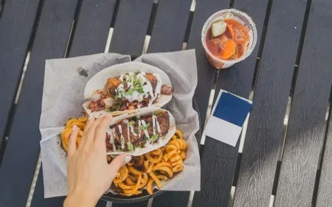 Hand reaching for tacos, curly fries, and a garnished drink.
