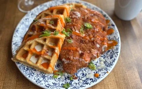 Plate of crispy chicken and waffles drizzled with sauces and green onions.