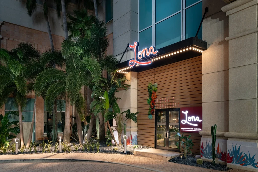 Modern Lona restaurant exterior at night with illuminated sign and tropical foliage.