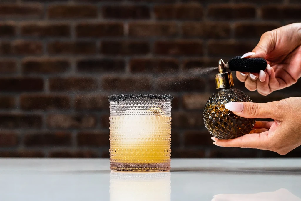 Hands spray atomizer mist over garnished cocktail on white surface.
