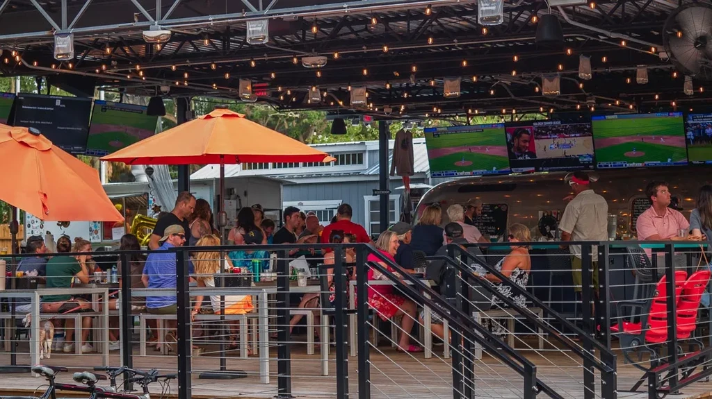 Bustling outdoor bar patio with Airstream, orange umbrellas, and sports TVs.