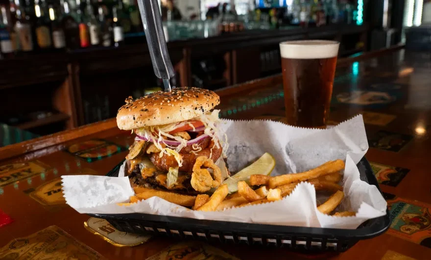 Stacked burger, fries, and dark beer on a wooden bar counter.