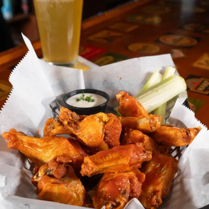 Basket of buffalo wings, celery, dipping sauce, and beer on a bar.