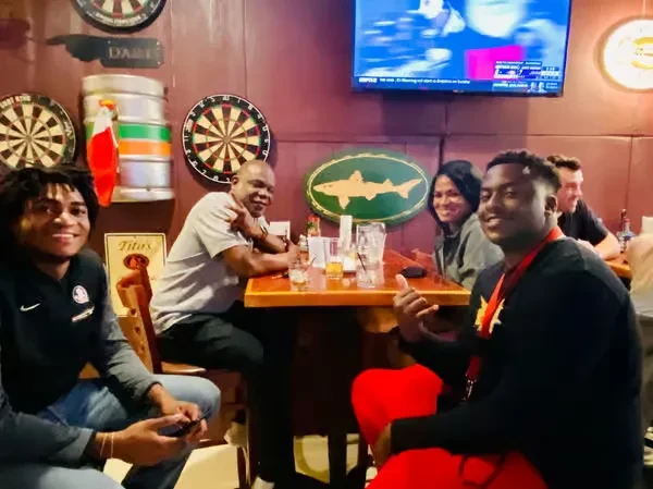 Smiling people drink and chat in a cozy pub with dartboards.