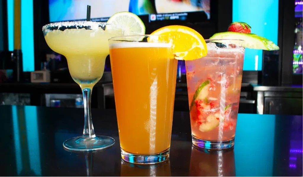 Margarita, amber drink, and strawberry cocktail on a bar with neon lights.