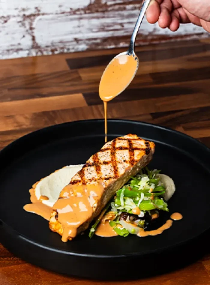 Hand drizzles sauce onto grilled salmon with purée and greens.
