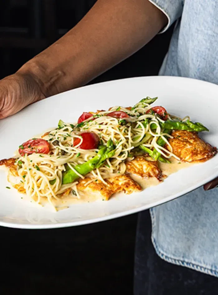 Hands holding plate of spaghetti with pan-seared fish, tomatoes, asparagus.