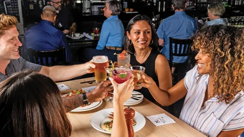 Four smiling people toast drinks at a lively restaurant table.