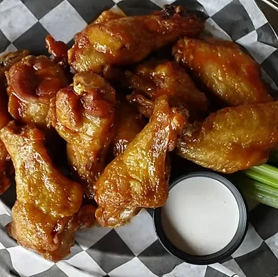 Piled, glossy chicken wings on checkered paper with dip and celery.