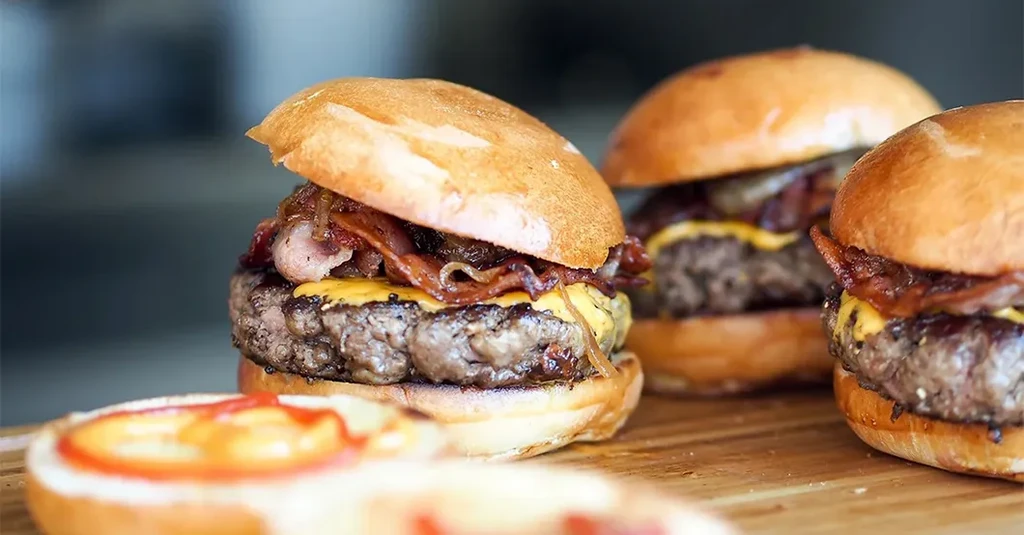 Multiple gourmet cheeseburgers with bacon, cheese, and onions on a wooden board.
