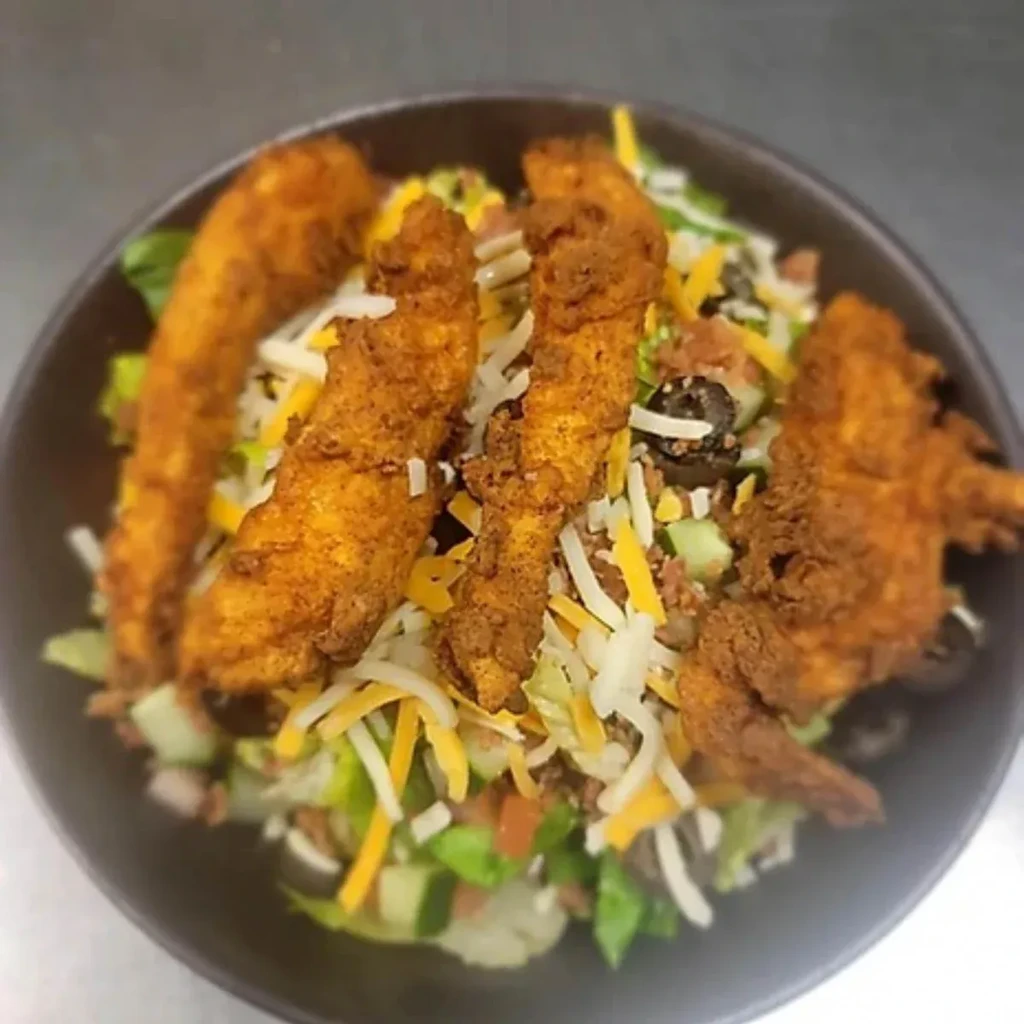 Salad with fried chicken tenders, cheese, olives, and bacon in a dark bowl.
