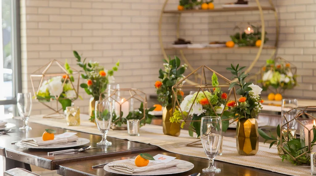 Elaborate long wooden table with gold terrariums, flowers, and orange fruit.