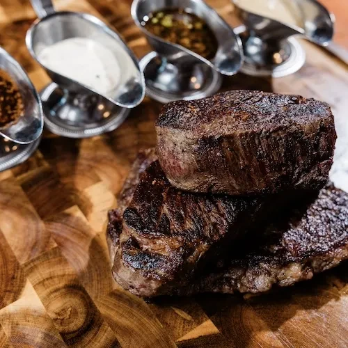Crispy seared steaks on wooden board, with four sauce boats.