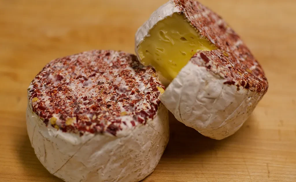 Two artisanal cheeses, one whole with red flakes, one cut, on wood.
