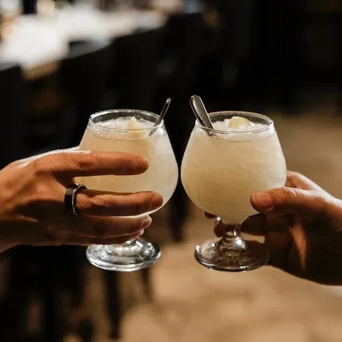Clinking hands hold stemware glasses with iced drinks and spoons.
