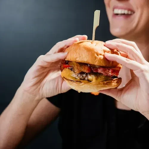 Close-up: Hands holding gourmet bacon burger, part of smiling face visible.