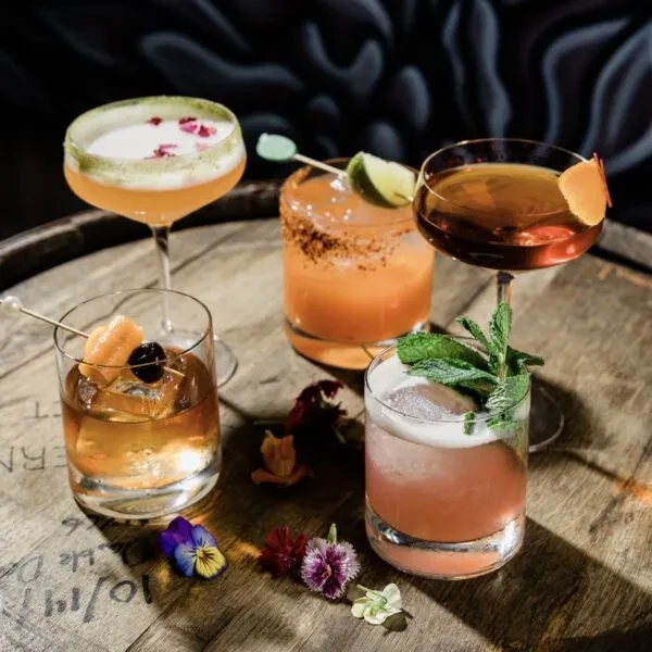 Five distinct cocktails with garnishes and edible flowers on a wooden surface.