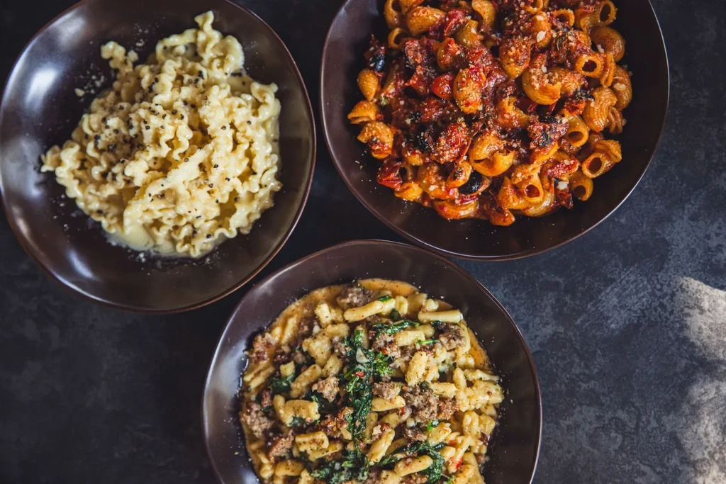 Overhead view of three colorful pasta dishes in dark serving bowls.