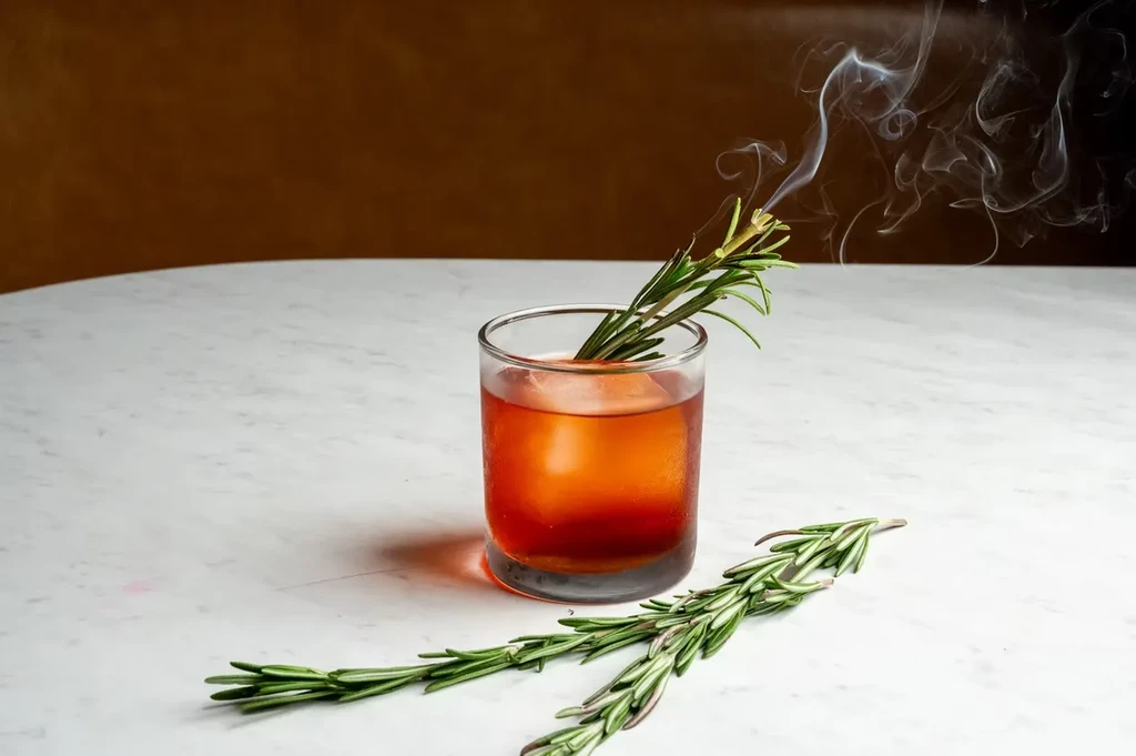 Amber cocktail in a short glass with smoking rosemary sprig.