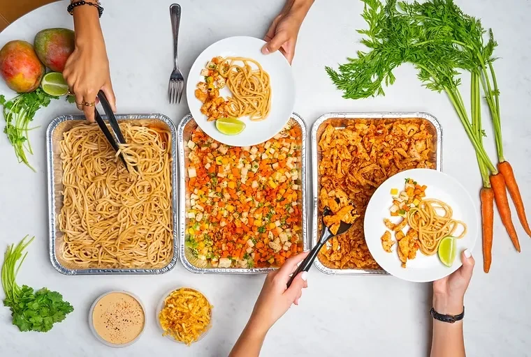 Hands serving noodles, vegetables, and seasoned chicken from trays.