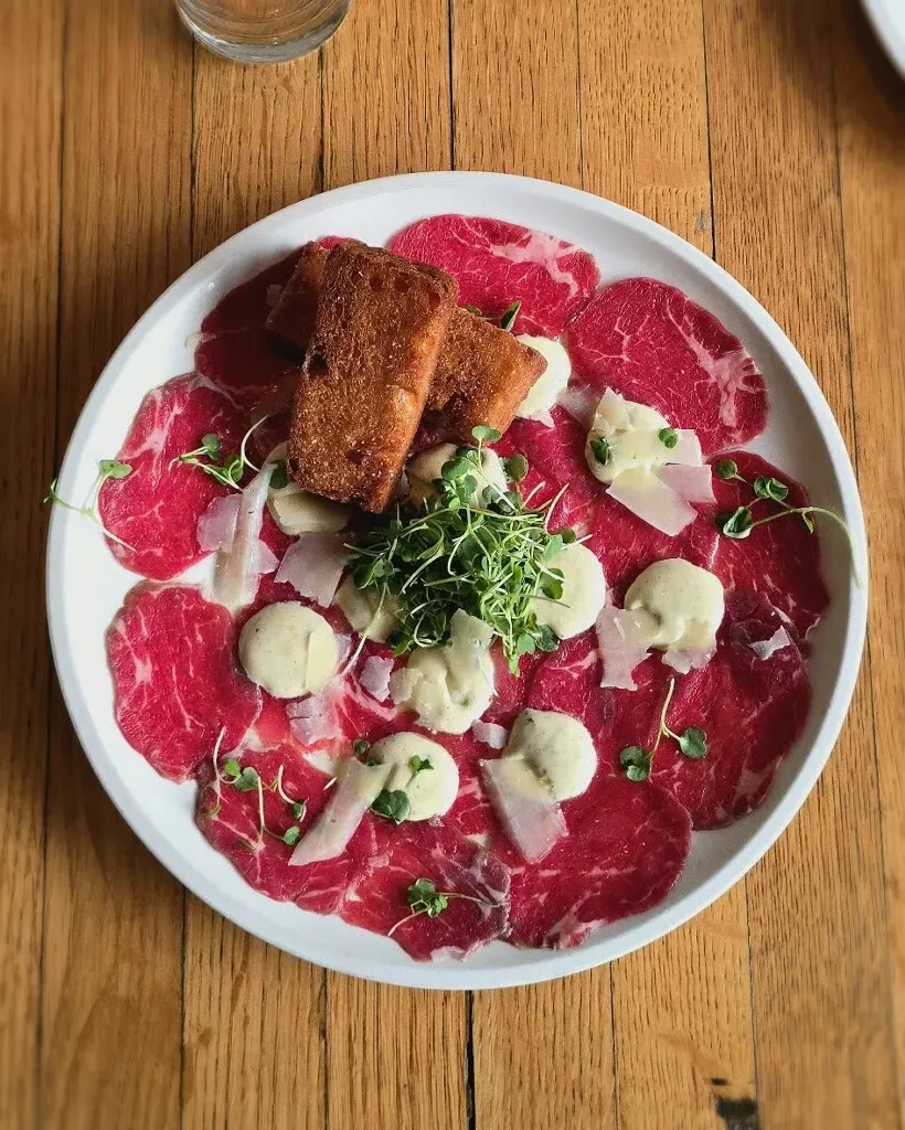 Dish of thinly sliced red meat carpaccio, parmesan, microgreens, and toasted bread.