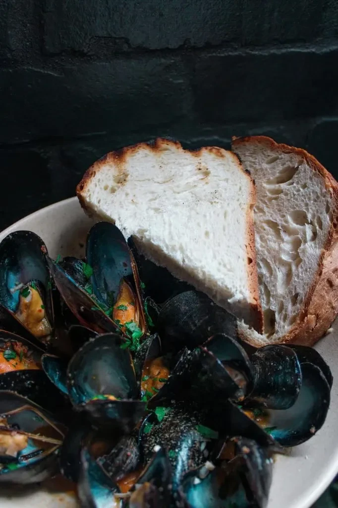 Dish of dark-shelled mussels in reddish broth, with rustic toasted bread.