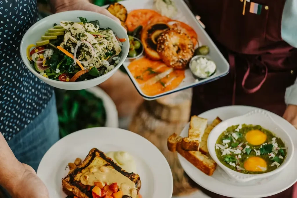 Hands presenting varied food dishes: salad, salmon, eggs, French toast.