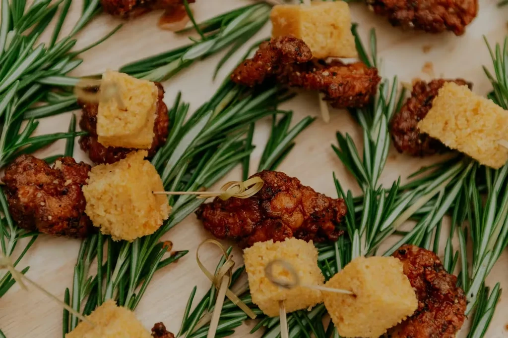 Glazed reddish-brown chicken, yellow cornbread skewers, and rosemary on wood.