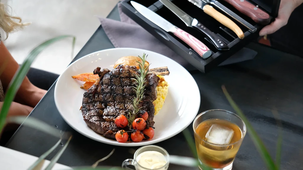 Grilled steak with sides, cocktail, and steak knives in a restaurant.