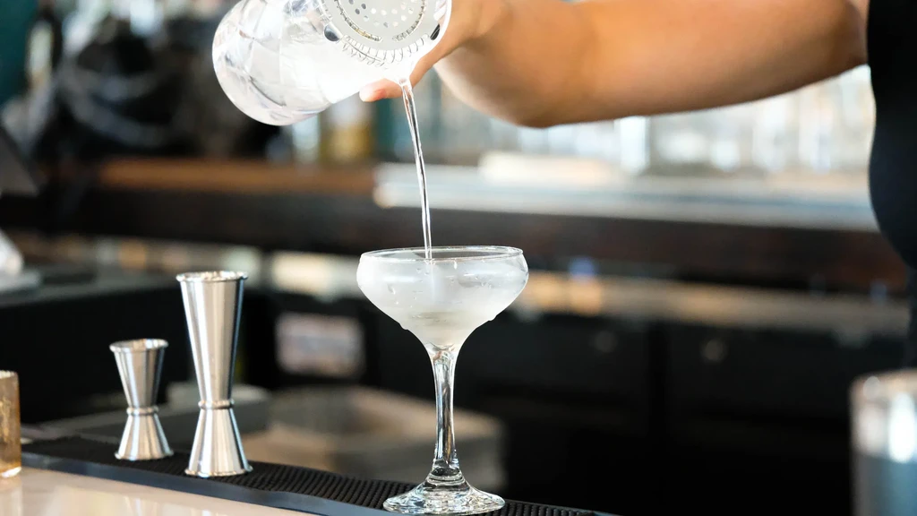 Bartender pours clear drink from shaker into iced coupe glass.