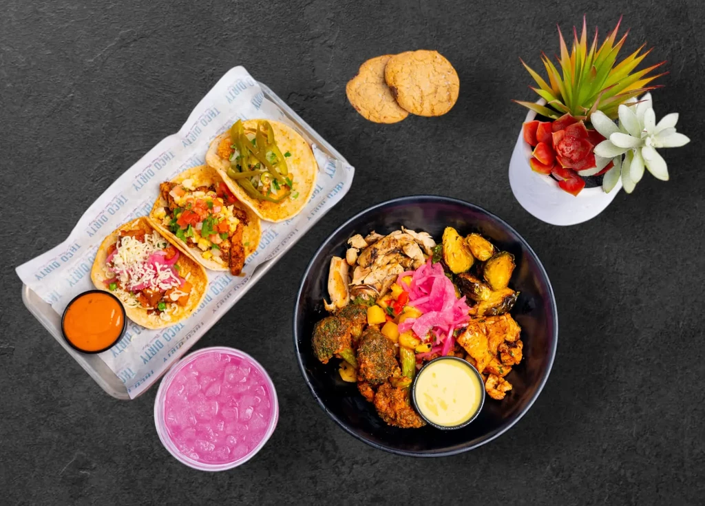 Overhead shot of tacos, a colorful protein bowl, pink drink, and cookies.