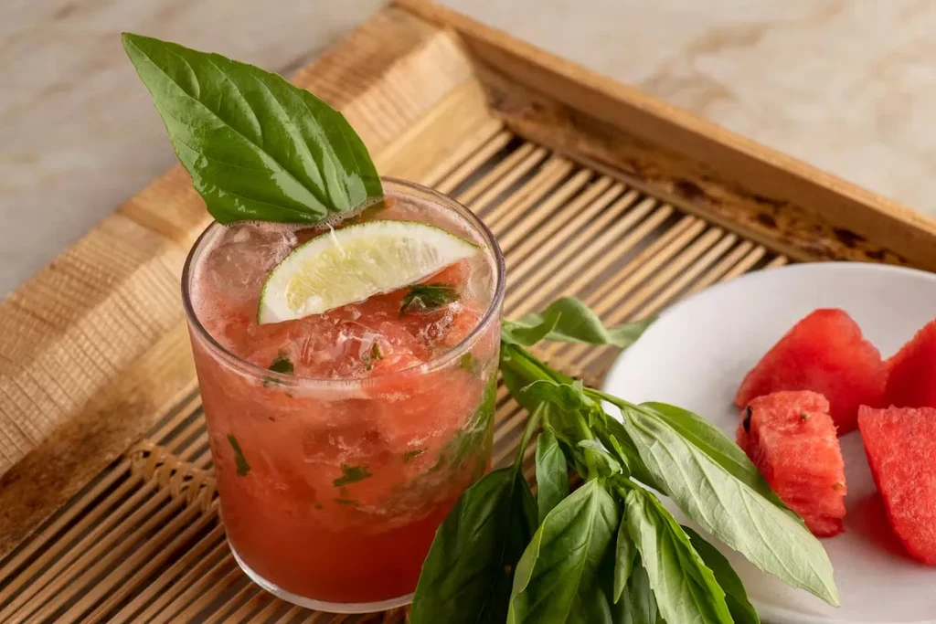 Iced red drink with lime, basil, and watermelon on a bamboo tray.