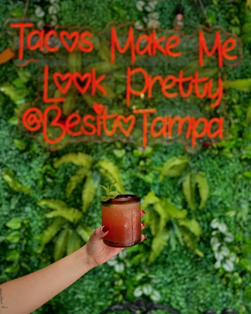 Hand holds red cocktail with mint; green wall, neon sign.